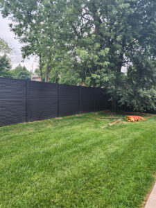 Neat, modern low-height dark grey WPC boundary fence separating a paved path from a well-maintained landscaped garden with shrubs and a white building in Germany.