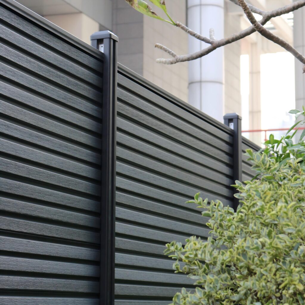 Modern dark grey composite fence with horizontal panels and aluminum posts, seamlessly integrated with green shrubs in urban outdoor setting.