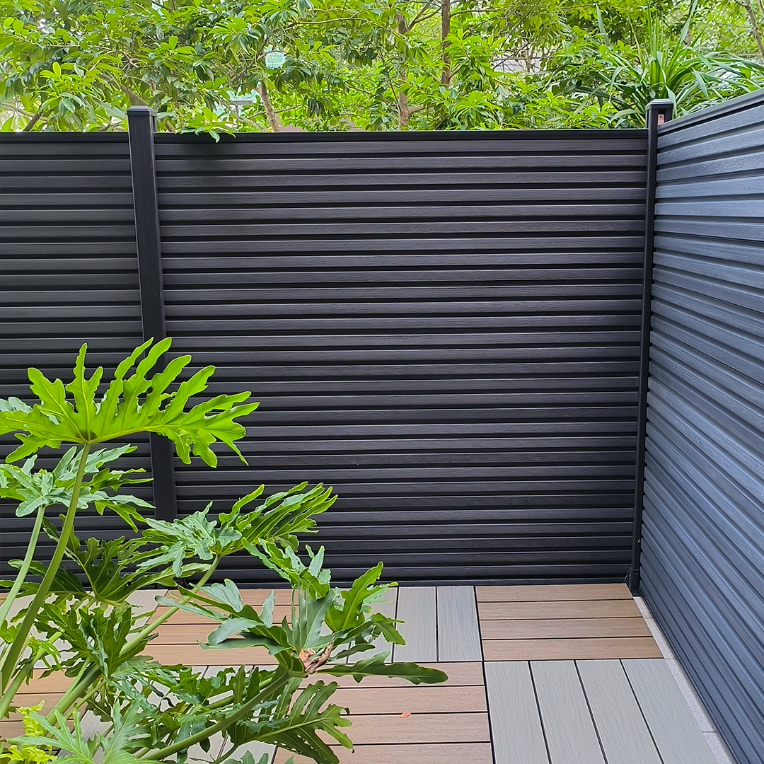 Mecofence dark grey horizontal composite fence with aluminum frame in horizontal arrangement, integrated with lush broad-leaf plants and light wood decking in modern outdoor setting