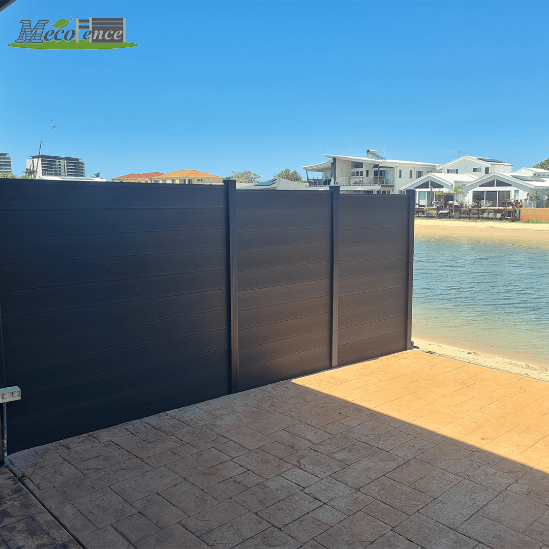 MecoFence 6ft Modern black composite privacy fencing panels installed next to a coastal shore with residential houses in the background.