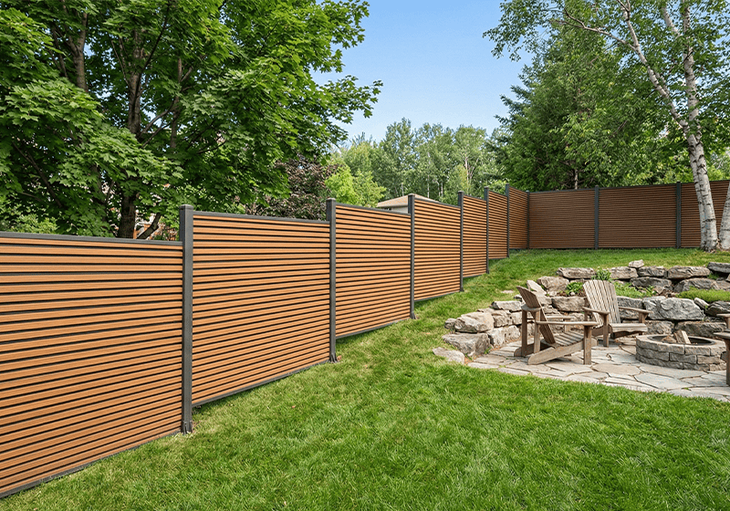 modern composite fence with horizontal brown slats and black posts installed in a Canadian backyard, featuring a stepped design on a slope, a stone patio, and mature trees.