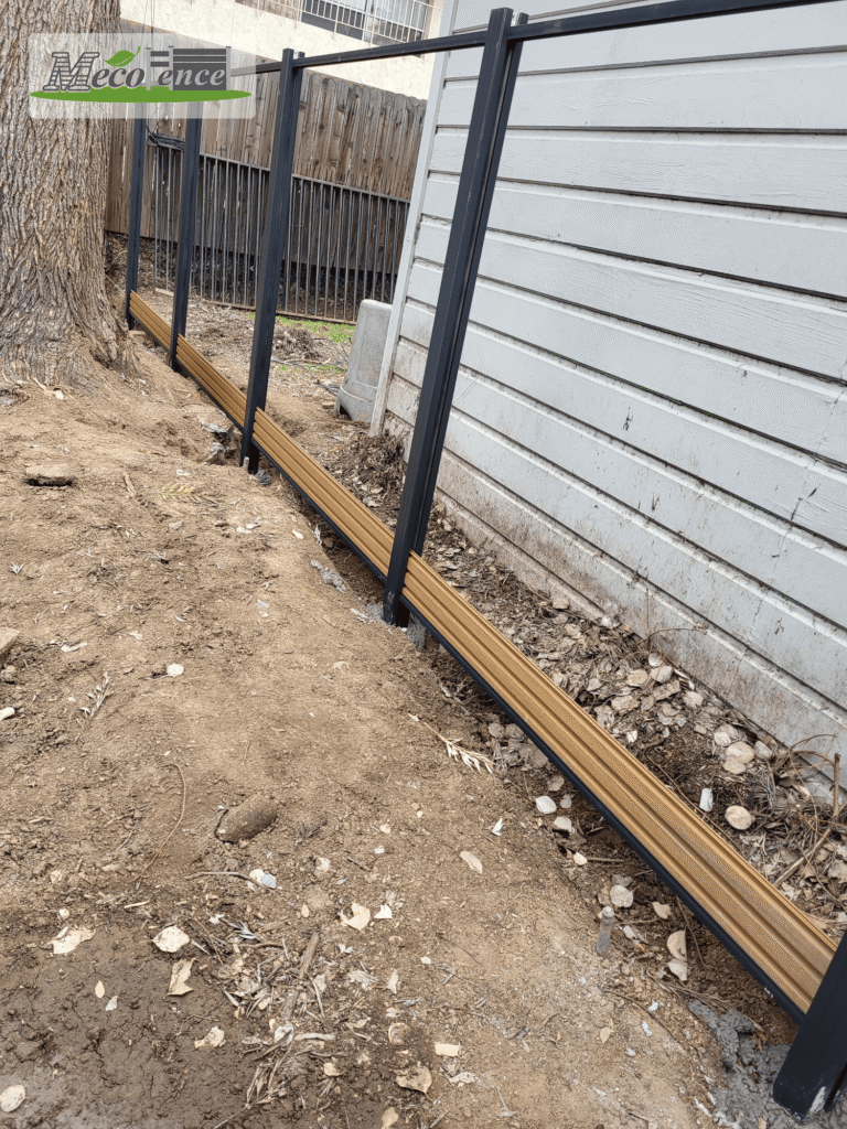Teak composite fence bottom rail installed on sloped grade against white siding, California residential install